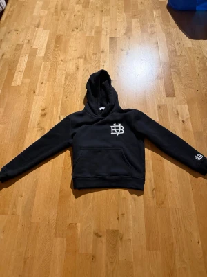 Svart hoodie från Bvaldi med logga - Svart hoodie från Bvaldi med stor huva och vit broderad logga på bröstet samt mindre logga på ärmen. Klassisk känguruficka framtill och mjukt material som känns skönt mot huden. Perfekt för en avslappnad och stilren look.