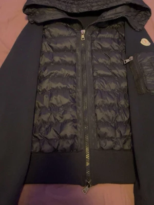 Svart dunjacka från Moncler - Snygg svart dunjacka från Moncler med quiltad front, huva och dragkedja. Jackan har en ficka med dragkedja på ärmen och Moncler-logga. Perfekt för kyliga dagar och ger en sportig look.