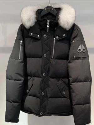 Svart dunjacka från Moose Knuckles - Snygg svart dunjacka från Moose Knuckles med stor fluffig vit pälskrage, silverfärgade dragkedjor och logga på ärmen. Jackan har flera fickor med dragkedja och tryckknappar, samt en quiltad design som ger en riktigt fet vinterlook.