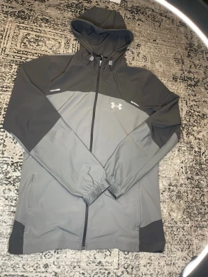  Under Armour Vindjacka  - Snygg tvåtonad grå vindjacka från Under Armour med huva och hel dragkedja framtill. Jackan har logga på bröstet, justerbar huva och elastiska muddar. Perfekt för blåsiga dagar och sportiga outfits.