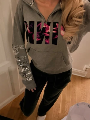 Grå PINK hoodie med paljetter - Snygg grå hoodie från Victoria's Secret PINK med stor blommig logga framtill och paljetttext på båda ärmarna. Halv dragkedja, magficka och ribbade muddar. Perfekt för en chill och trendig look.