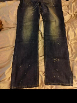 Dsq2  blå bootcut jeans - Säljer ett par dsq2  blå jeans med bootcut passform. Jeansen har slitna detaljer och ljusa partier framtill och baktill, samt vita stänk för en trendig look. Klassisk femficksmodell i kraftigt bomullsjeansmaterial.