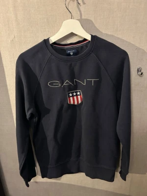 Mörkblå sweatshirt från GANT - Mörkblå sweatshirt från GANT med broderad logga och amerikanskinspirerat sköldmärke på bröstet. Tröjan har rund halsringning, långa ärmar och ribbade muddar. Perfekt för en avslappnad och stilren look.