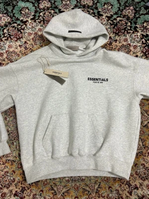 Grå Essentials Fear of God hoodie - Snygg ljusgrå hoodie från Essentials Fear of God med svart logga på bröstet och ryggen. Klassisk känguruficka framtill och huva. Perfekt för en chill och trendig look.