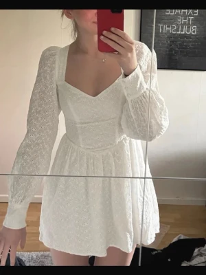 Vit kort klänning med öppen rygg - Superfin vit kort klänning med broderade detaljer och långärmade ärmar. Klänningen har v-ringning framtill och öppen rygg med knytband. Perfekt för student eller sommaren. Inga defekter och bara använd en gång.
