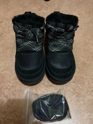 Ugg lowmel skor - Svarta kängor från UGG med grov sula och snörning. Ovandel i mocka och textil, fodrad med mjuk päls. Skorna har rund tå och extra skosnören medföljer. Perfekta för kyliga dagar och har en stilren, sportig look.