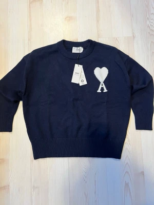 Mörkblå stickad tröja från Ami Paris - Mörkblå stickad tröja från Ami Paris med rund halsringning och ribbade muddar. På bröstet finns ett vitt hjärta och bokstaven A, som är Ami Paris signaturdetalj. Tröjan har en avslappnad passform och är tillverkad i mjukt material.