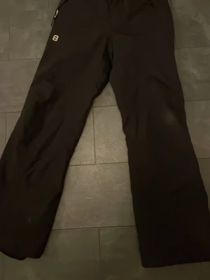 Svarta täckbyxor från 8848 Altitude - Svarta täckbyxor från 8848 Altitude med dragkedja och knapp framtill. Byxorna har en enkel design med en liten logga på vänster lår och praktiska fickor med dragkedja. Perfekta för kalla dagar och vintersport.