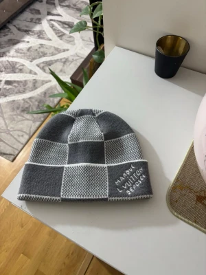 Grå mössa  - Snygg grå och vit rutig mössa från  med broderad logga framtill. Mössan är stickad i mjukt material och har en klassisk passform som passar perfekt till kyliga dagar.