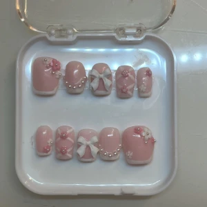 Rosa och vita nageldekorationer - Söta nageldekorationer i rosa och vitt med pärlor och blommor. Setet innehåller naglar med små rosetter och blommor, perfekt för en gullig och elegant look. Kommer i en praktisk förvaringsask.