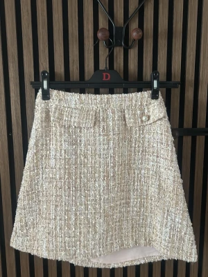Beige kortkjol i tweed med knappdetalj - Stilren beige kortkjol i tweed med diskret glittrig yta och en dekorativ knapp framtill. Kjolen har en lätt A-linjeform och är fodrad för extra komfort. Perfekt för dig som gillar en chic och trendig look.