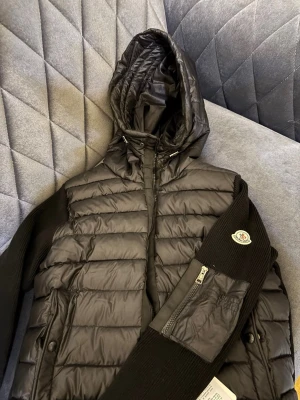 Svart dunjacka med huva från Moncler - Snygg svart dunjacka från Moncler med quiltad front, stickade ärmar och huva. Jackan har dragkedja framtill, fickor med dragkedja och en Moncler-logga på ärmen. Perfekt för kyliga dagar när du vill vara både varm och stilren.