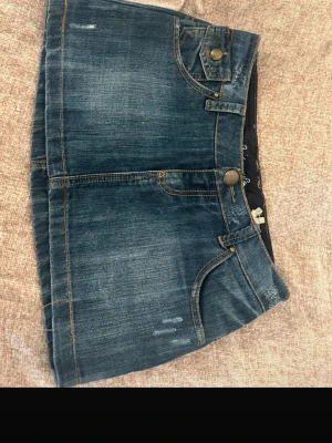 Blå jeanskjol med fickor - Snygg blå jeanskjol med klassiska fickor fram och bak, samt dekorativa sömmar och detaljer i beige på bakfickorna. Kjolen har knapp och dragkedja framtill och är i en kort modell. Perfekt för en trendig look. Funkar super i liten S storlek också🩷 köpte den secondhand, men har aldrig kunna använda den själv,
