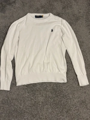 Vit stickad tröja från Polo Ralph Lauren - Klassisk vit stickad tröja från Polo Ralph Lauren med rund halsringning och diskret broderad logga på bröstet. Tröjan har ribbade muddar vid ärmslut och nederkant, vilket ger en snygg och stilren look. 