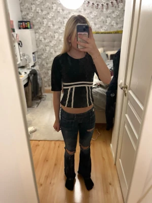 Svart croppad topp med öppen rygg - Snygg svart croppad topp med vita detaljer och öppen rygg med knyt. Toppen har korta ärmar och en unik design framtill med vita linjer. Perfekt för dig som vill sticka ut med en trendig och cool look.