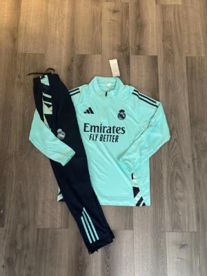 Real Madrid träningsset Adidas mint/svart - Snyggt Real Madrid träningsset från Adidas med mintgrön långärmad tröja och svarta byxor med mintgröna detaljer. Tröjan har dragkedja vid halsen och klubbmärke på bröstet. Byxorna har resår i midjan, klubbmärke och Adidas-logga. Produkten är M i junior