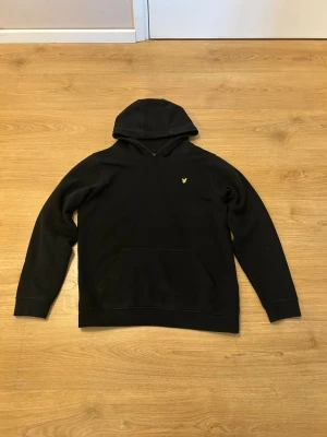 Lyle&Scott Hoodie - NYPRIS: 800kr   Svart hoodie från Lyle & Scott Junior med klassisk gul logga på bröstet. Hoodien har en stor känguruficka framtill och en mjuk huva. Tillverkad i bomullsmix som känns skön mot huden. Perfekt för en avslappnad och stilren look.