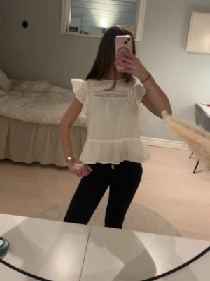 Vit blus med volang och spetsdetaljer - Söt vit blus med volang nertill och spetsdetaljer upptill. Blusen har korta, voluminösa ärmar och en luftig passform som ger en romantisk vibe. Perfekt att styla med jeans eller kjol för en trendig look. Fortfarande prislappen kvar 