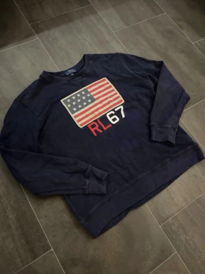 Polo Ralph Lauren crewneck - Mörkblå sweatshirt från Polo Ralph Lauren med amerikansk flagga och RL67 tryck på bröstet. Perfekt för en avslappnad och stilren look. Storlek XL med passar L‼️