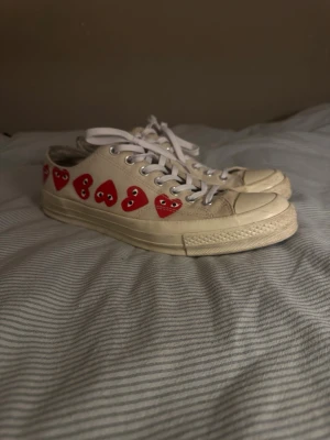Converse x CDG Play vita sneakers - Snygga Converse x Comme des Garçons Play sneakers i vitt canvas med röda hjärtan på sidorna. Klassisk låg modell med snörning och rund tå. Perfekta för dig som vill sticka ut med en ikonisk design och coola detaljer.