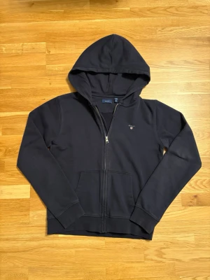 Marinblå zip hoodie från Gant - Snygg marinblå hoodie från Gant med dragkedja framtill och två fickor. Klassisk huva och diskret Gant-logga broderad på bröstet. Tillverkad i mjukt bomullsmaterial som känns skönt mot huden. Perfekt för en avslappnad och stilren look.