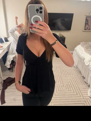 Svart v-ringad topp med kort ärm - Svart topp med djup v-ringning och korta ärmar. Modellen har en figurnära passform upptill och faller lösare nedtill. Perfekt att styla med jeans eller kjol för en trendig look.