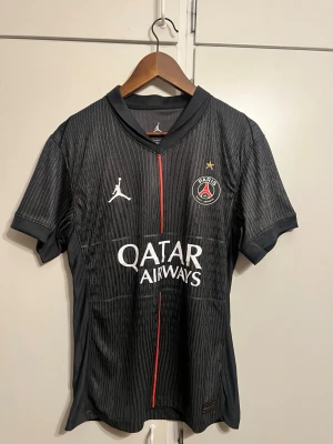 Svart PSG matchtröja Jordan - Snygg svart PSG matchtröja med tunna vertikala ränder, röd detalj i mitten och klubbmärke på bröstet. Perfekt för fotboll eller träning. Passar S/M.