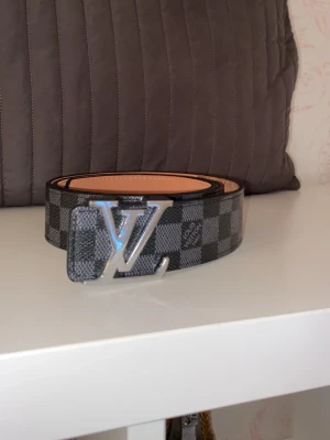 L-bälte svart/grå - Snyggt bälte i svart och grått damier-mönster med stor logga i silver som spänne. Bältet är gjort i läder och har en klassisk, stilren look som passar till jeans eller kostymbyxor.