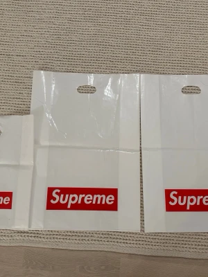 Supreme shoppingväska i plast - Vit shoppingväska från Supreme med det klassiska röda Supreme-logotypen tryckt på framsidan. Väskan är tillverkad i plast och har utskurna handtag för enkel bärning. Perfekt för dig som gillar streetwear och vill ha en ikonisk accessoar.