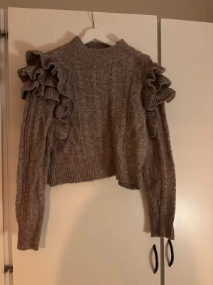 Brun stickad tröja med volangärmar - Supersnygg brun stickad tröja med höga volangdetaljer på axlarna och långa ärmar. Tröjan har en ribbad, hög hals och är croppad i modellen. Perfekt för dig som vill ha något extra till din outfit. Materialet känns mjukt och mysigt.