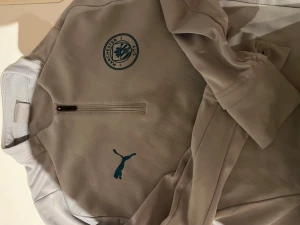Grå Manchester City träningströja Puma - Snygg grå träningströja från Puma med Manchester Citys klubbmärke på bröstet. Tröjan har halv dragkedja framtill och är tillverkad i ett mjukt, funktionellt material. Perfekt för träning eller uppvärmning.