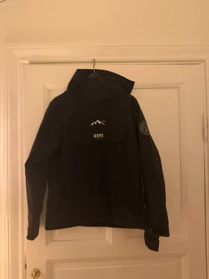 Svart vindjacka från Dope med huva - Säljer en svart vindjacka från Dope med stor huva och praktisk magficka med logga och bergmotiv. Jackan har lång ärm, dragsko i huvan och patch på ena ärmen. Perfekt för dig som vill ha en stilren och funktionell jacka. Den är endast använd ett fåtal gånger 