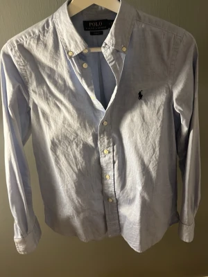 Ljusblå skjorta från Polo Ralph Lauren - Klassisk ljusblå skjorta från Polo Ralph Lauren med button-down krage och broderad logga på bröstet. Skjortan har långa ärmar, knappar framtill och är tillverkad i mjuk bomull. Perfekt för en stilren look.