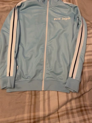 Ljusblå ziptröja från Palm Angels - Snygg ljusblå ziptröja från Palm Angels med vita och svarta ränder längs ärmarna. Tröjan har hög krage, dragkedja framtill och ribbade muddar. Perfekt för en sportig och trendig look. Materialet är troligtvis polyester.