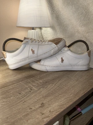 Vita och guldiga sneakers från Polo Ralph Lauren -  Obs köpte av en annan på plick,Snygga vita sneakers från Polo Ralph Lauren med guldiga detaljer på tå, häl och plös. Klassisk låg modell med snörning och Polo-logga på sidan. Tillverkade i skinn för en clean och stilren look.