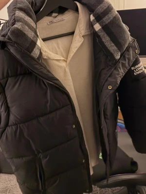 Svart pufferjacka från Burberry - Snygg svart pufferjacka från Burberry med klassiskt rutigt foder i huvan och logga på ärmen. Jackan har dragkedja framtill och är quiltad för extra värme. Perfekt för kalla dagar och riktigt stilren look.