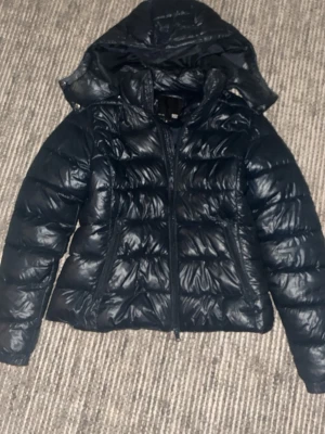 Svart dunjacka med huva - Snygg svart dunjacka med quiltad design och huva. Jackan har dragkedja framtill och en klassisk pufferlook som håller dig varm under kalla dagar. Perfekt för dig som vill ha en stilren och funktionell jacka till vintern.