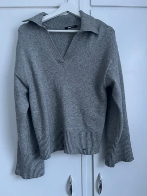 Grå stickad tröja med krage - Säljer en stilren grå stickad tröja från GINA TRICOT. Tröjan har en bred krage, djup V-ringning och ribbstickade muddar vid ärmslut och nederkant. Perfekt för lager på lager och enkel att matcha med jeans eller kjol.
