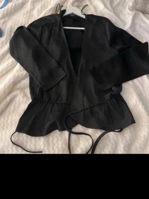Svart omlottblus med knytband - Oanvänd!     Snygg svart omlottblus med långa ärmar och knytband i midjan. Blusen har en lätt glansig finish och volangdetalj nedtill som ger en trendig look. Perfekt att styla med jeans eller kjol för en chic outfit.