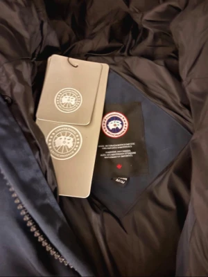 Mörkblå dunjacka från Canada Goose - Snygg mörkblå dunjacka från Canada Goose med huva och klassisk patch på ärmen. Jackan har quiltad design, dragkedja framtill och två stora fickor. Perfekt för kalla dagar och riktigt varm. Passar dig som vill ha både stil och funktion.