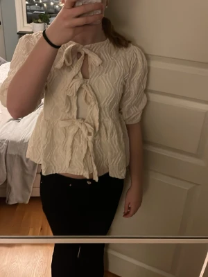 Krämvit blus med knyt och struktur - Supersöt krämvit blus med tre dekorativa knyt framtill och puffiga trekvartsärmar. Blusen har ett vågigt, strukturerat tyg som ger en unik look och en lätt utsvängd passform. Perfekt för dig som gillar trendiga detaljer och romantisk stil.