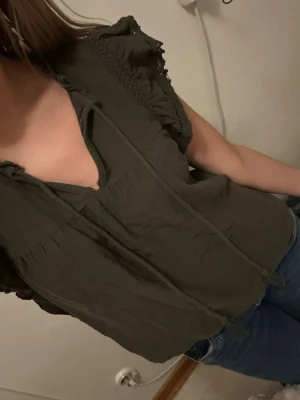 Mörkgrön blus med volang och knyt - Supersnygg mörkgrön blus från Moss Copenhagen med volangdetaljer på axlarna och knytband soppar vid halsen. Blusen är ärmlös och har en avslappnad passform, perfekt till jeans eller kjol. Materialet känns lätt och luftigt.