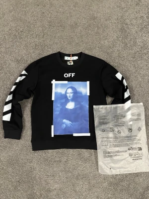 Off-White svart sweatshirt Mona Lisa - Svart sweatshirt från Off-White med vit text 'OFF' och blå Mona Lisa-print framtill. Vita pilar på ryggen och diagonala ränder på ärmarna. Rund hals och ribbade muddar. Trendig streetwear med ikonisk design.