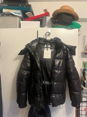 Svart Moncler Maya dunjacka med huva - Snygg svart dunjacka från Moncler med glansig finish och stor huva. Jackan har quiltad design, dragkedja framtill och Moncler-logga på ärmen. Perfekt för kalla dagar och ger en riktigt trendig look.
