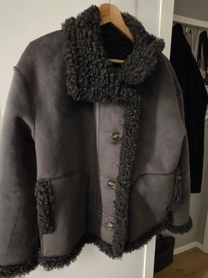Mörkgrå teddyjacka med fårpälslook - Säljer en mörkgrå teddyjacka med fårpälslook och stora krage. Jackan har knappar framtill, två fickor och detaljer i pälsimitation. Perfekt för kalla dagar och ger en riktigt mysig vibe.