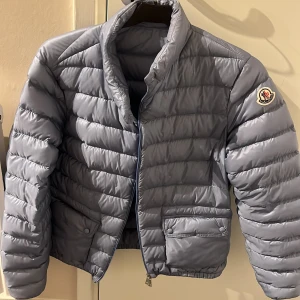 Ljusblå  LANS  från Moncler - Säljer en snygg ljusblå äkta dunjacka från Moncler med klassisk quiltad design och Moncler-logga på ärmen. Jackan har dragkedja framtill, två fickor med lock och knapp samt hög krage. Perfekt för kyliga dagar. Storlek 3 . Liten i storlek . Köpt på Wakkakuu mer som en mellan  jacka 
