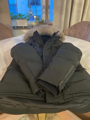 Svart parkas från Moose Knuckles - Snygg svart parkas från Moose Knuckles med pälskantad huva och flera praktiska fickor. Jackan har dragkedja och knappar framtill samt ribbade muddar vid ärmsluten. Perfekt för kalla dagar med sin varma och rejäla design.