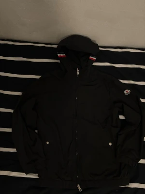 Svart vindjacka från Moncler - Svart vindjacka från Moncler med huva och coola röd, vit och blå detaljer på huvan. Jackan har dragkedja framtill, två fickor med tryckknappar och Moncler-logga på ärmen. Materialet är lätt och syntetiskt, perfekt för blåsiga dagar. Hör av dig om du har några frågor. Inga defekter. Behöver pengar vill göra mig av med den snabbt. Snabb affär 500kr. Har redan sänkt mycke mer än vad jag ville. 