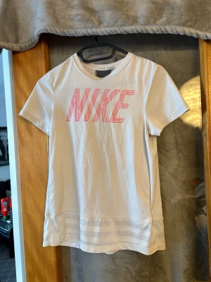 Vit Nike t-shirt med rosa tryck - Snygg vit t-shirt från Nike med stort rosa logotyptryck framtill och en grå vertikal rand på ryggen. Klassisk passform och rund halsringning. Tillverkad i mjuk bomull som känns skön mot huden. Perfekt för en sportig och avslappnad stil.