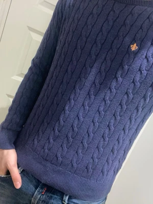 Mörkblå kabelstickad tröja Morris - Kabelstickad mörkblå tröja från Morris med rund halsringning och ribbade muddar. Tröjan har ett broderat gult emblem på bröstet och är tillverkad i mjukt material. Perfekt för en stilren och klassisk look.